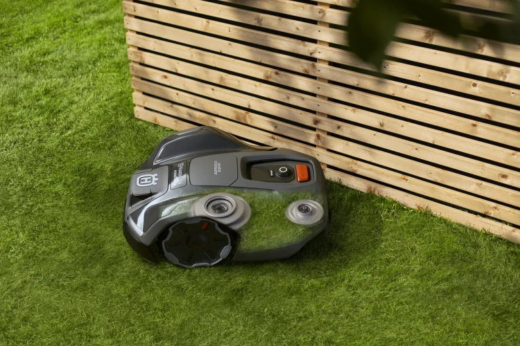 Robot koszący Husqvarna Automower® 405XE NERA w kolorze czarnym, z widocznymi kółkami oraz przyciskiem start. Produkt leży na trawie, blisko drewnianego ogrodzenia. Powierzchnia jest gładka, a logo Husqvarna jest czytelne.