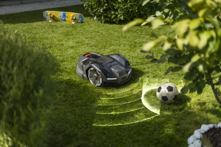 Robot koszący Husqvarna Automower® 405XE NERA w czarnej matowej obudowie na trawie, widoczny z boku z przyciskiem i kółkami. Obok piłka nożna, w tle skateboard i zieleń w naturalnym świetle.