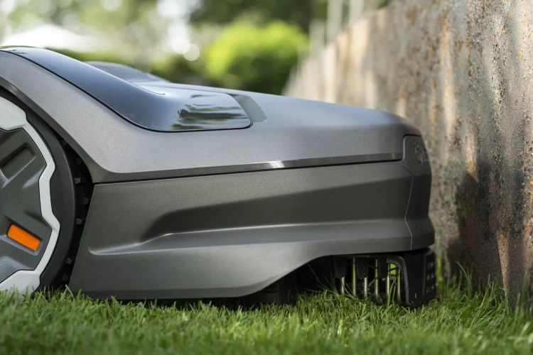 Robot koszący Husqvarna Automower® 405XE NERA w kolorze szarym, widoczny z boku, przylegający do murka. Widać detale takie jak czarne kółka, białe akcenty i pomarańczowy przycisk. Tło to trawnik oraz naturalny krajobraz w tle.