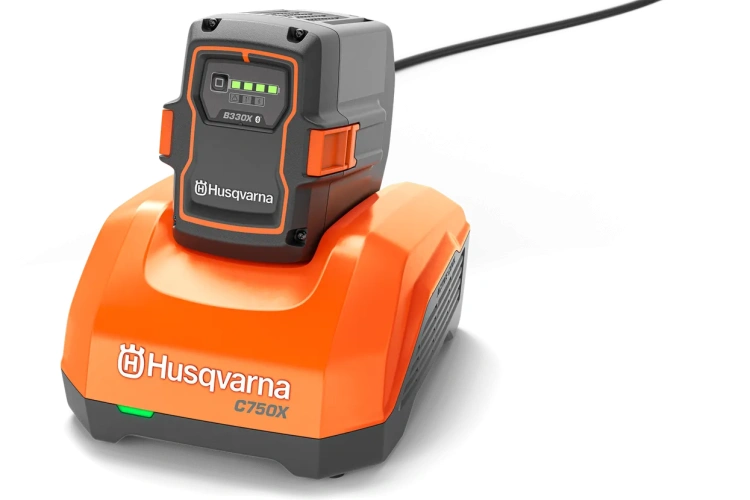Ładowarka Husqvarna 40-C750X w pomarańczowej obudowie z czarnym panelem i ekranem LED. Leży na powierzchni, kabel podłączony z tyłu, logo Husqvarna na froncie.
