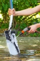 Pistolet zraszający Gardena w szaro-pomarańczowej kolorystyce, trzymany w ręku, skierowany na łopatę pokrytą ziemią. Produkt w akcji, na tle zieleni, w pozycji poziomej. Woda wypływa z końcówki, efektywnie usuwając brud.