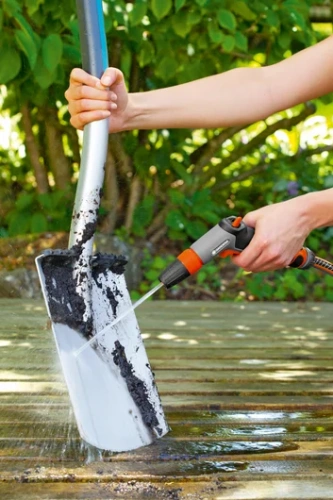 Pistolet zraszający Gardena w szaro-pomarańczowej kolorystyce, trzymany w ręku, skierowany na łopatę pokrytą ziemią. Produkt w akcji, na tle zieleni, w pozycji poziomej. Woda wypływa z końcówki, efektywnie usuwając brud.