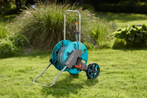 Wózek na wąż Gardena CleverRoll M w błękicie, z metalową konstrukcją, stabilną podstawą i kółkami. Ujęcie z boku pokazuje rolki i uchwyt do transportu, stoi na zielonej trawie.