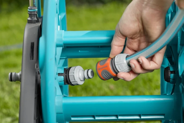 Błękitny wózek na wąż CleverRoll M Easy Gardena na trawie, stabilny. Ręka podłącza wąż z pomarańczową złączką do białego adaptera. Widok z boku w trakcie użycia.