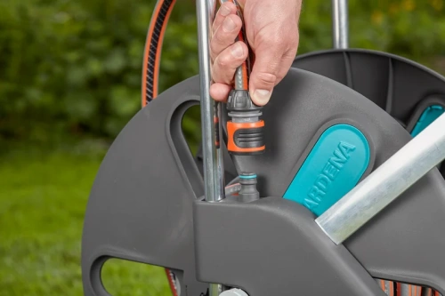 Wózek na wąż CleverRoll L Easy Gardena w szaro-niebiesko-pomarańczowej kolorystyce z systemem zwijania i złączem, używany na zewnątrz w ogrodzie, z ręką mocującą wąż do uchwytu.