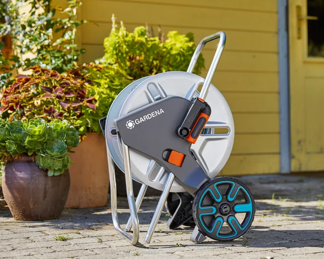 Zestaw wózka na wąż GARDENA CleverRoll M Metal w szaro-białym kolorze, stojący, z metalową ramą i niebieskimi kołami. Widoczne logo GARDENA oraz uchwyt do przenoszenia. Tło: kwiaty w donicach, przy domowym wejściu.