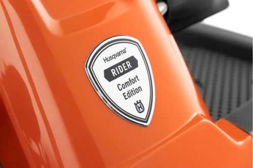Logo Husqvarna Rider Comfort Edition na pomarańczowej obudowie. Emblemat w kształcie tarczy z białym tłem i czarnymi napisami, detal i kolorystyka wyraźnie widoczne na neutralnym tle.