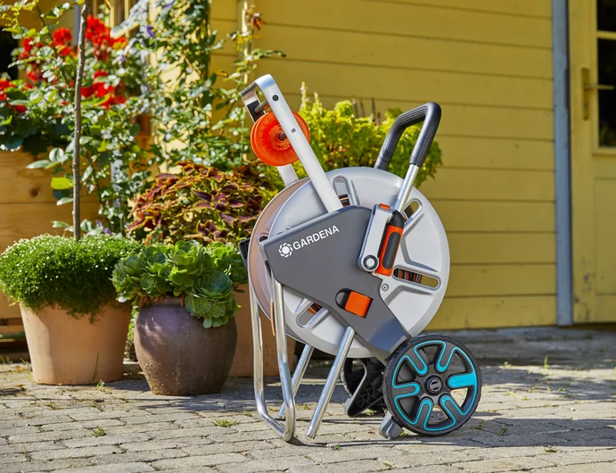 Zestaw wózka na wąż CleverRoll M Metal GARDENA w kolorze szarym i pomarańczowym, stojący, z metalową konstrukcją, zamontowany na kółkach. Widoczne logo GARDENA oraz uchwyt do transportu. Tło z kwiatami i drewnianą ścianą domu.