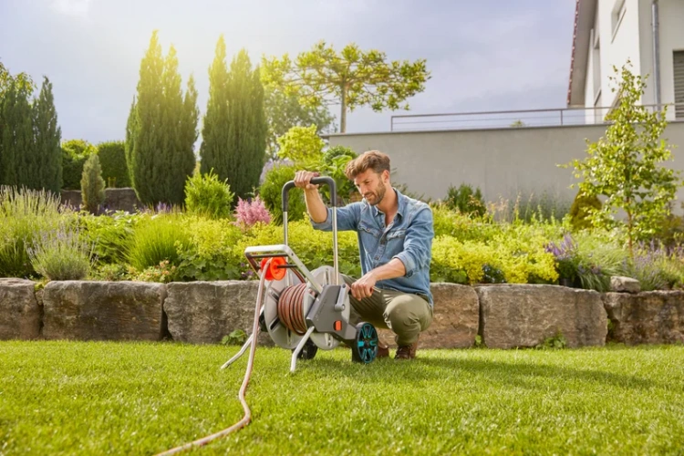 Zestaw wózka na wąż CleverRoll M Metal GARDENA w kolorze szarym z wbudowanym bębnem i czerwonym wężem, stojący na trawie. Mężczyzna pochyla się nad wózkiem, sprawdzając mechanizm. Tło to zadbany ogród z roślinami i drzewami.