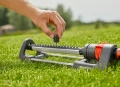 Zraszacz wahadłowy Aqua M GARDENA ulokowany na trawie, z ręką ustawiającą kontrolę przepływu wody. Produkt w kolorze szaro-czerwonym, z metalowym korpusem i plastikowymi elementami. Ujęcie z boku, pokazujące detale konstrukcji.