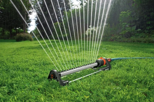 Zraszacz wahadłowy Aqua L GARDENA na zielonym trawniku, z rozpryskującą się wodą. Plastikowy, szaro-czarny z pomarańczowymi elementami, leżący na tle drzew i krzewów.