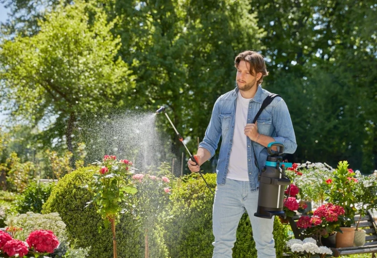 Opryskiwacz ciśnieniowy Gardena Plus 5L trzymany przez mężczyznę w ogrodzie. Produkt o szarym korpusie z niebieskimi detalami, z wężem i dyszą w ręku, spryskuje kwiaty. Zgromadzone rośliny tworzą zielone tło z kolorowymi kwiatami w tle.