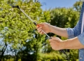 Opryskiwacz plecakowy Gardena z teleskopową końcówką, trzymany w dłoni. Metalowa rurka z regulacją sprysku, czarna końcówka, pomarańczowy uchwyt. W tle zieleń drzew, ujęcie z boku, ukazujące czynność użycia.