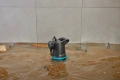 Pompa zanurzeniowa do brudnej wody Gardena w kolorze czarnym z niebieskimi akcentami, umieszczona w wodzie z liśćmi i plastikowymi butelkami. Produkt widoczny z boku w otoczeniu zanieczyszczonego środowiska.