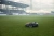 Automower® 580 EPOS® Husqvarna w kolorze szaro-pomarańczowym, stojący na trawie na stadionie. W zbliżeniu widoczny kształt urządzenia z widocznymi kołami oraz otworami wentylacyjnymi. Tło to murawa z trybunami w lekkiej mgle.