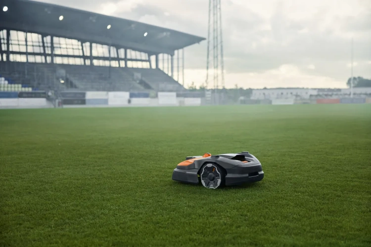 Automower® 580 EPOS® Husqvarna w kolorze szaro-pomarańczowym, stojący na trawie na stadionie. W zbliżeniu widoczny kształt urządzenia z widocznymi kołami oraz otworami wentylacyjnymi. Tło to murawa z trybunami w lekkiej mgle.