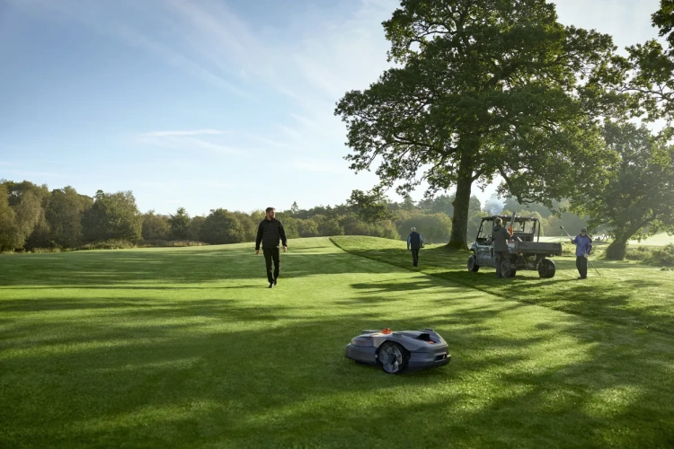 Automower® 580L EPOS® Husqvarna na zielonej trawie, z przodu po prawej stronie kadru, z mężczyzną w czarnym stroju spacerującym w tle. Robot koszący o szarej obudowie i pomarańczowym znaku rozpoznawczym, widoczny w naturalnym otoczeniu.