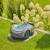 Robot koszący Gardena Sileno SMART 1200 na trawniku, szary z niebieskimi detalami, z panelem sterującym i pomarańczowym przyciskiem. Tło to bujna zieleń i biało-kremowe kwiaty, podkreślające ogród.