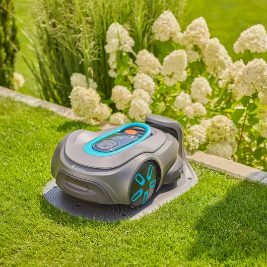 Robot koszący Gardena Sileno SMART 1200 na trawniku, szary z niebieskimi detalami, z panelem sterującym i pomarańczowym przyciskiem. Tło to bujna zieleń i biało-kremowe kwiaty, podkreślające ogród.