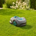 Robot koszący Gardena Sileno SMART Free 600 w kolorze szarym z niebieskimi akcentami, umieszczony na trawniku. Ujęcie z boku, ukazujące gładką, zaokrągloną obudowę oraz panel sterowania na górze. W tle widoczne zielone rośliny i białe kwiaty.