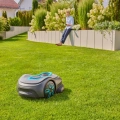 Robot koszący Gardena Sileno SMART Free 600 w szaro-niebieskich barwach na trawniku z kwiatami. W tle osoba na ławce steruje robotem za pomocą urządzenia.