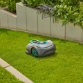 Robot koszący Gardena Sileno SMART Free 1200 w kolorze szarym z niebieskimi akcentami, w pozycji leżącej na trawie. Ujęcie z boku, ukazujące kształt oraz detale obudowy. W tle widoczna zielona roślinność i betonowe obrzeża ogrodu.