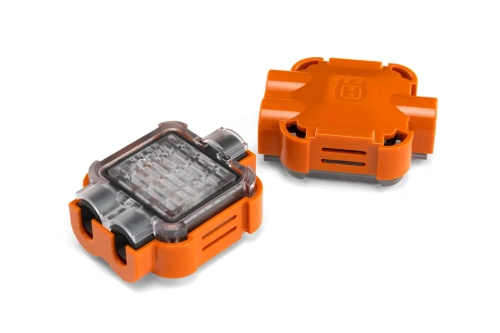 Pomarańczowy łącznik przewodu Automower Husqvarna z przezroczystą częścią, gładki, zaokrąglony, z otworami do mocowania i logo, na białym tle, widoczny z boku, wykonany z tworzywa sztucznego.