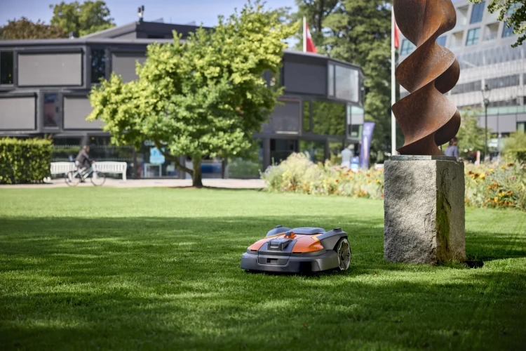 Robot koszący Husqvarna Automower® 560 Epos w kolorze czarno-pomarańczowym, widoczny na zielonym trawnikiu. Ujęcie z boku, sprzęt z bliska, częściowo zatrzymany obok kamiennego postumentu. W tle budynek oraz zieleń drzew.