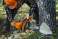 Pilarka Husqvarna 445S z pomarańczowym korpusem i czarną rączką, cięcie pnia drzewa. Użytkownik w kasku i odzieży roboczej, na tle zielonej trawy i drzew.