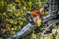 Pomarańczowa pilarka Husqvarna 445S z metalową prowadnicą, trzymana w rękawicach, przycina gałęzie na tle żółtozielonych liści, z widocznym logo i przełącznikami.