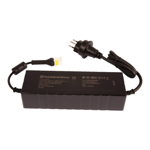 Zasilacz robota koszącego Automower 310E NERA Husqvarna w kolorze czarnym, prostokątny, z szeregami żeber na bokach. Widoczny kabel z wtyczką oraz złącze do robota. Logo Husqvarna na górze. Produkt ułożony leżąco na jednolitym tle.