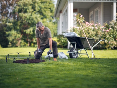 Mężczyzna w szarym T-shircie klęczy na trawie, tworząc obszar wymaganego montażu dla zestawu ogrodzeniowego. Obok widoczny wózek ogrodowy, łopata i worek z ziemią. W tle widać dom z ogrodem pełnym róż.