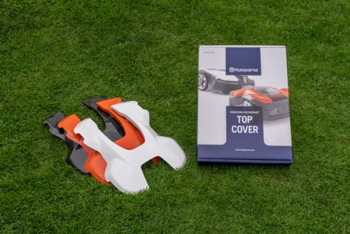 Obudowa białego i pomarańczowego robota koszącego Husqvarna AUTOMOWER 105 na trawie, obok instrukcja z grafiką robota w prostokątnej teczce. Prosta aranżacja eksponuje urządzenie i dokumentację.