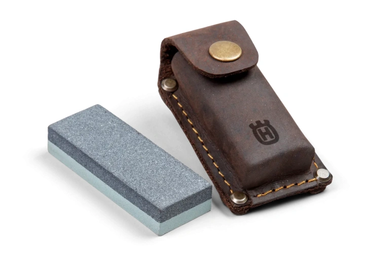 Osełka do ostrzenia siekier Husqvarna w kolorze szarym  Produkt ujęty z frontowej pozycji, ukazujący osełkę w kształcie prostokąta w odcieniu szarym. Obok widoczny futerał w kolorze brązowym o strukturze skóry i z widocznym logotypem Husqvarna