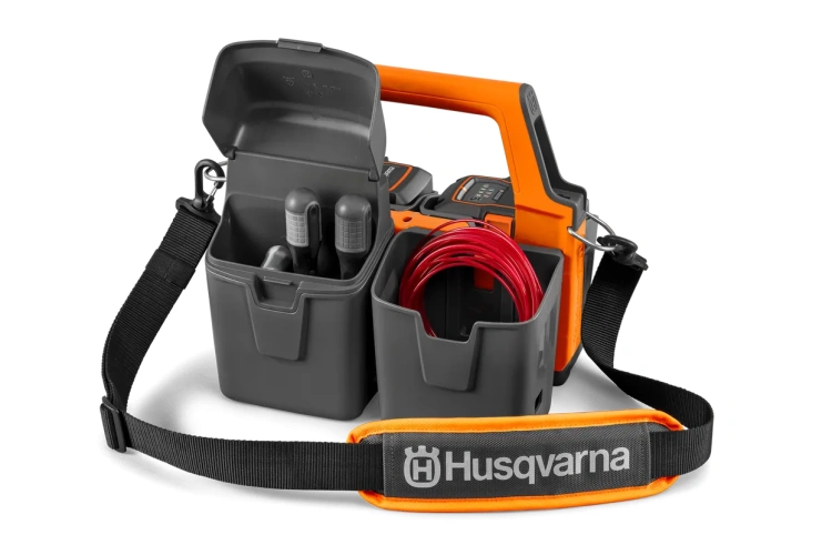 zasobnik na akumulatory do pilarki Husqvarna w kolorze szaro-pomarańczowym. Z przodu widoczne są zbiorniki na akumulatory.  zamontowane w praktycznej torbie z uchwytem. Logo Husqvarna na pasku ułatwia transport.