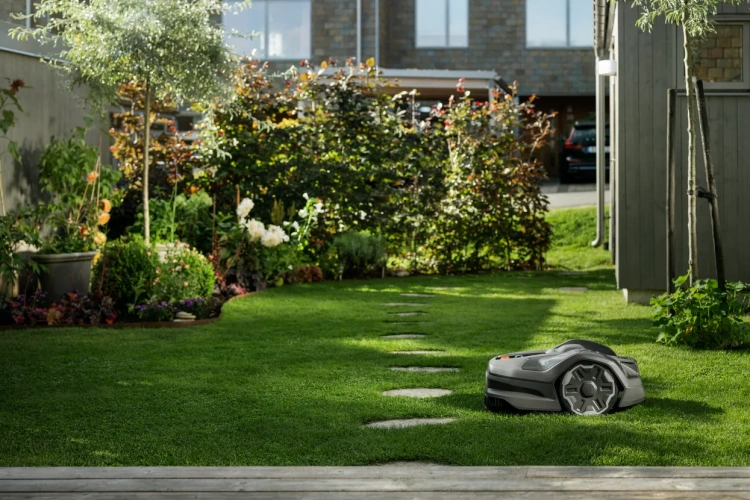 Automower® 305E NERA, robot koszący Husqvarna, w kolorze grafitowym. Ujęty podczas pracy w ogrodzie. Zobacz z jaką dokładnością kosi trawę. Robot koszący pokazany z ujęcia bocznego.