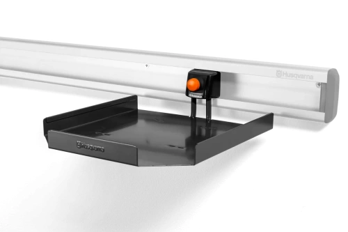 Półka do przechowywania akumulatorów i ładowarek Husqvarna, wykonana z tworzywa o prostokątnym płaskim kształcie w kolorze szarym ciemny. Produkt prezentowany z boku z widocznym mocowaniem do montażu na ścianie. Na białym tle.