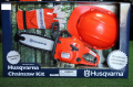 Zestaw Husqvarna: pomarańczowa zabawkowa pilarka z metalowym łańcuchem i logo, czarny uchwyt, biało-czarne rękawice leżące obok oraz kask po boku na ciemnym tle.