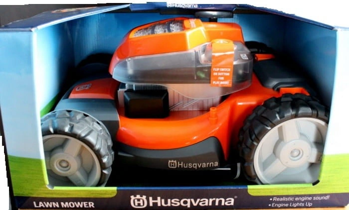 Zabawka kosiarka AWD Husqvarna w pomarańczowym kolorze z szarymi detalami, na kolorowym opakowaniu. Ma przełącznik, teksturowaną podstawę oraz realistyczne detale, jak światło silnika i logo Husqvarna.