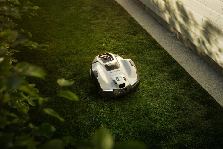 Robot koszący Husqvarna Automower® 410VE NERA w białej matowej obudowie na trawie, widoczny z boku z przyciskiem i kółkami. Obok piłka nożna, w tle  zieleń w naturalnym świetle.