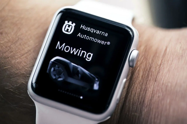 Wyświetlacz smartwatcha z logotypem Husqvarna Automower, na którym widoczny jest napis Mowing. Obudowa w kolorze srebrnym, z białym paskiem. Tło głębokorobione wskazuje, że produkt jest w użyciu na nadgarstku.