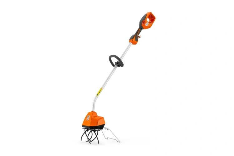 Kultywator TH125i Husqvarna w kolorze pomarańczowym z czarnym silnikiem. Ujęcie z boku, stojący. Wyposażony w metalowe ostrza, jednostka z uchwytem i kontrolami na górze. Przeznaczony do pracy w ogrodzie. Tło jednolite.