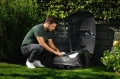 Domek dla ropbota koszącego wraz z roboterm koszącym automower przy którym człowiek dotyka robota koszącego
