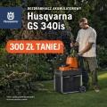 Rozdrabniacz akumulatorowy Husqvarna GS 340is w kolorze szarym z pomarańczowym akcentem, stojący na trawie, trzymany przez użytkownika. Widać koła i rączkę do transportu, a w tle kwitnące rośliny, co sugeruje zastosowanie w ogrodzie.