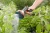 Premium pistolet zraszający wielofunkcyjny Gardena z ergonomicznym uchwytem i okrągłą końcówką. Ujęcie z boku, w tle widać kwiaty oraz drzewka. Kobieta prowadzi rynnę do podlewania roślin.