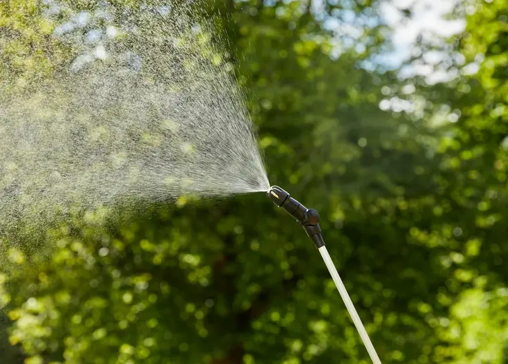 Opryskiwacz ciśnieniowy Gardena EasyPump 5L z metalową rurką i czarną dyszą. Uchwyt z pomarańczowym przyciskiem. Trzymany pionowo na tle drzew, używany w ogrodzie.