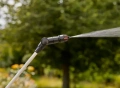 Opryskiwacz ciśnieniowy Gardena EasyPump 5L z metalową rurką i czarną dyszą. Uchwyt z pomarańczowym przyciskiem. Trzymany pionowo na tle drzew, używany w ogrodzie.