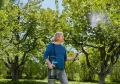 Opryskiwacz ciśnieniowy Gardena EasyPump 5L trzymany przez mężczyznę w ogrodzie. Produkt o szarym korpusie z niebieskimi detalami, z wężem i dyszą w ręku, spryskuje kwiaty. Zgromadzone rośliny tworzą zielone tło z kolorowymi kwiatami w tle.