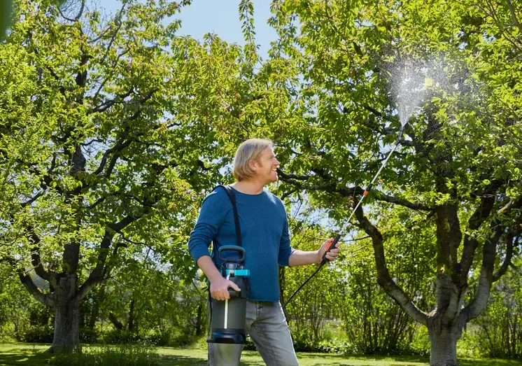 Opryskiwacz ciśnieniowy Gardena EasyPump 5L trzymany przez mężczyznę w ogrodzie. Produkt o szarym korpusie z niebieskimi detalami, z wężem i dyszą w ręku, spryskuje kwiaty. Zgromadzone rośliny tworzą zielone tło z kolorowymi kwiatami w tle.