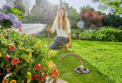 Puszka poboru wody 8266-20 Gardena, czarna z pomarańczowym złączem, na trawie. Zbliżenie na rękę używającą węża ogrodowego do podłączenia produktu. W tle kwiaty i zielona przestrzeń ogrodowa.